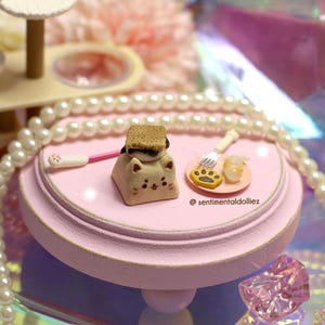 K&ouml;nnte beinhalten: Eine Miniatur-Katzenfigur mit einem S'more darauf, einem rosa Serviertablett und einem kleinen Teller mit einem Pfotenabdruck-Keks. Die Figur hat ein niedliches Gesicht und ist von einer Perlenkette umgeben. Der Text "@sentimentaldolliez" ist sichtbar.