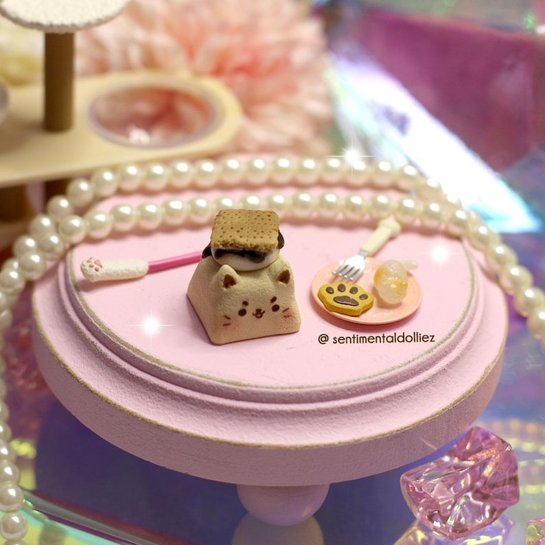 K&ouml;nnte beinhalten: Eine Miniatur-Katzenfigur mit einem S'more darauf, einem rosa Serviertablett und einem kleinen Teller mit einem Pfotenabdruck-Keks. Die Figur hat ein niedliches Gesicht und ist von einer Perlenkette umgeben. Der Text "@sentimentaldolliez" ist sichtbar.