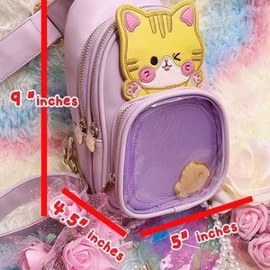 K&ouml;nnte beinhalten: Eine lavendelfarbene Kunstleder-Umh&auml;ngetasche mit einer durchsichtigen Plastiktasche auf der Vorderseite. Die Tasche hat eine gelbe Katzenapplikation mit rosa Ohren und einen goldenen Kettenriemen. Die Tasche ist 23 cm hoch und 13 cm breit. Der Riemen ist 11 cm lang.