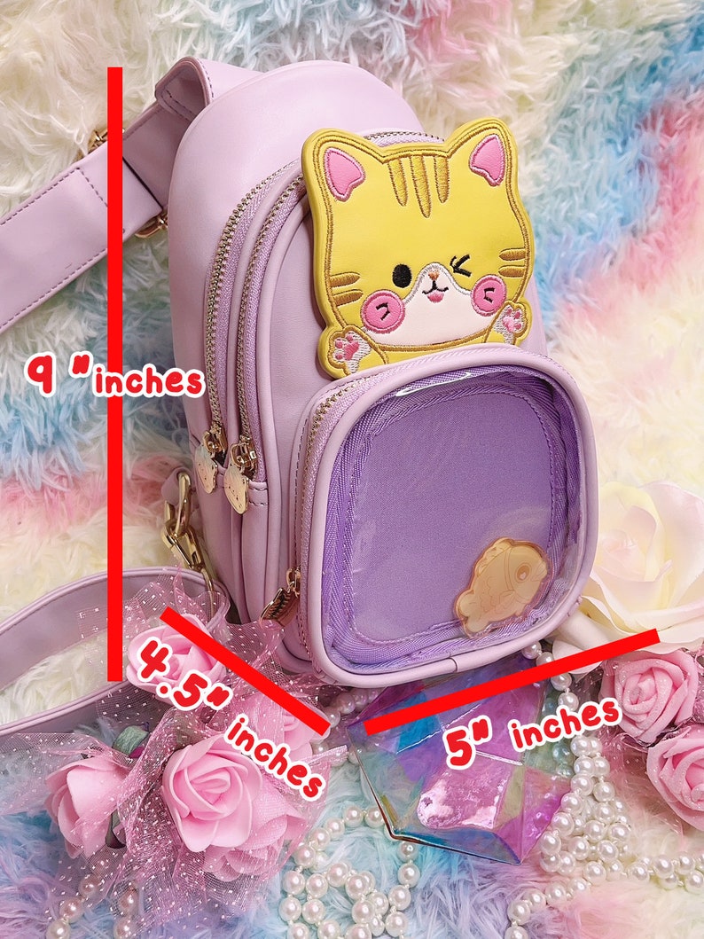 K&ouml;nnte beinhalten: Eine lavendelfarbene Kunstleder-Umh&auml;ngetasche mit einer durchsichtigen Plastiktasche auf der Vorderseite. Die Tasche hat eine gelbe Katzenapplikation mit rosa Ohren und einen goldenen Kettenriemen. Die Tasche ist 23 cm hoch und 13 cm breit. Der Riemen ist 11 cm lang.