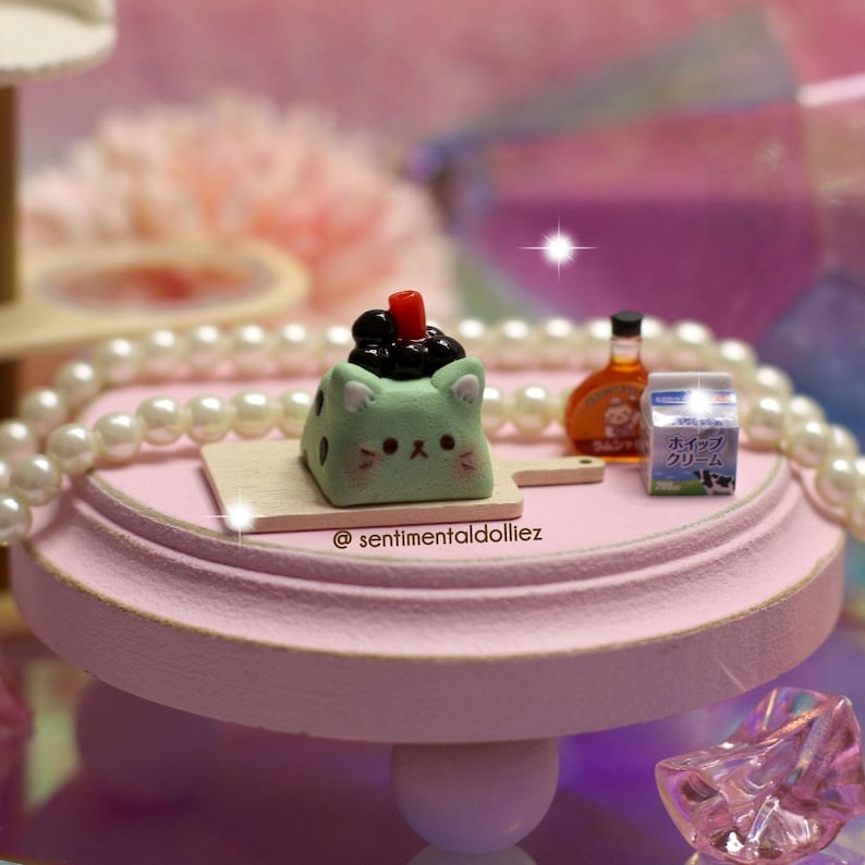 May include: A miniature, light green cat-shaped dessert with black toppings and a red garnish sits on a wooden board. Beside it are a small bottle and a carton of milk. The scene is set on a pink, tiered stand, with a pearl necklace and text that reads "@sentimentaldolliez".