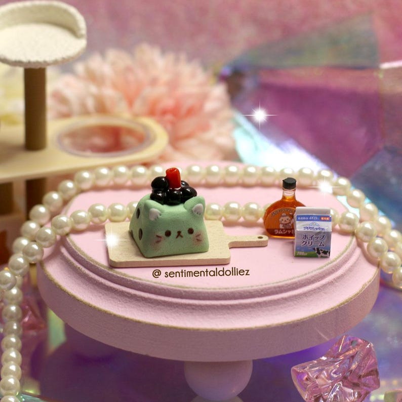 May include: A miniature scene featuring a light green cat-shaped dessert with black toppings on a wooden cutting board. Also, a small bottle and carton of cream are on a pink stand, with a pearl necklace and a cat tree in the background.