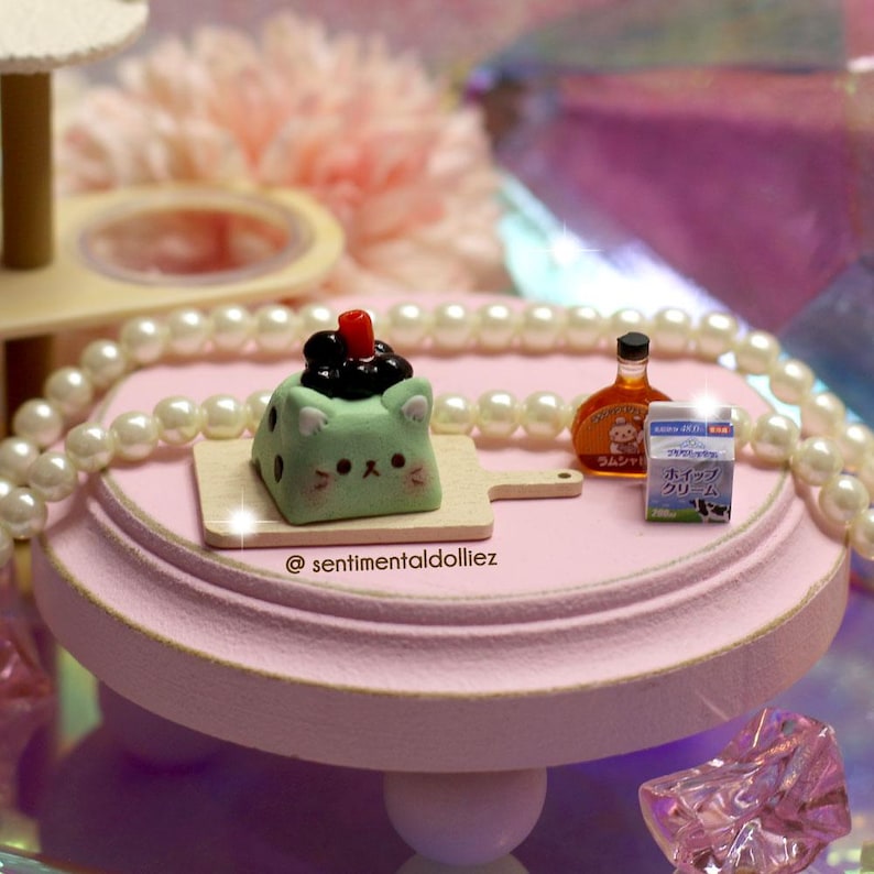 May include: A miniature scene featuring a light green cat-shaped dessert with black toppings and a red cherry on a small wooden cutting board. Also present are a small bottle of liquor and a carton of Japanese cream, all on a pink, tiered stand.