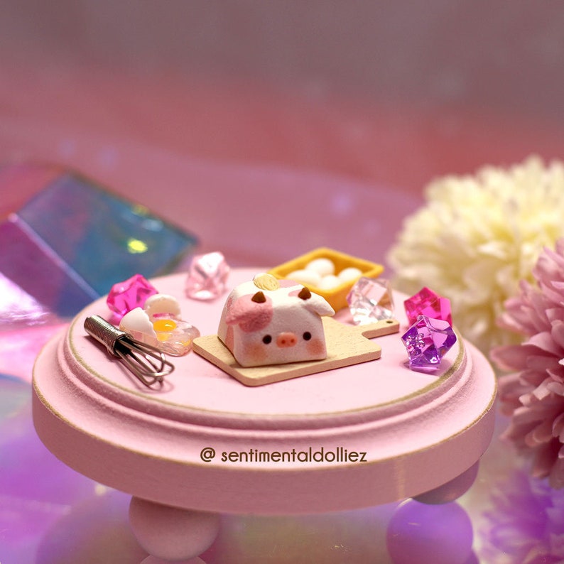 May include: A miniature pink pig-shaped cake on a wooden cutting board with a pink whisk, a broken egg, and a small basket of eggs on a pink pedestal. The scene is decorated with pink and purple gemstones.