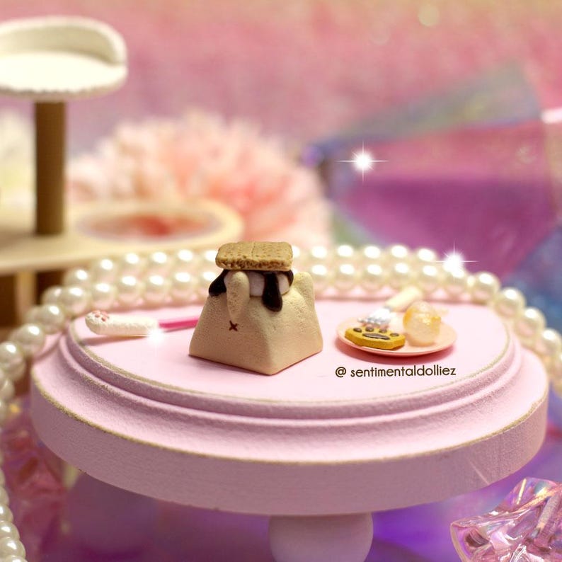 K&ouml;nnte beinhalten: Eine Miniatur-Dessertszene mit einem S'more auf einem rosa Sockeltisch. Der S'more hat einen Keks oben, Marshmallow und Schokolade. Ein kleiner Teller mit Miniatur-Desserts befindet sich in der N&auml;he. Der Tisch ist von Perlen umgeben.
