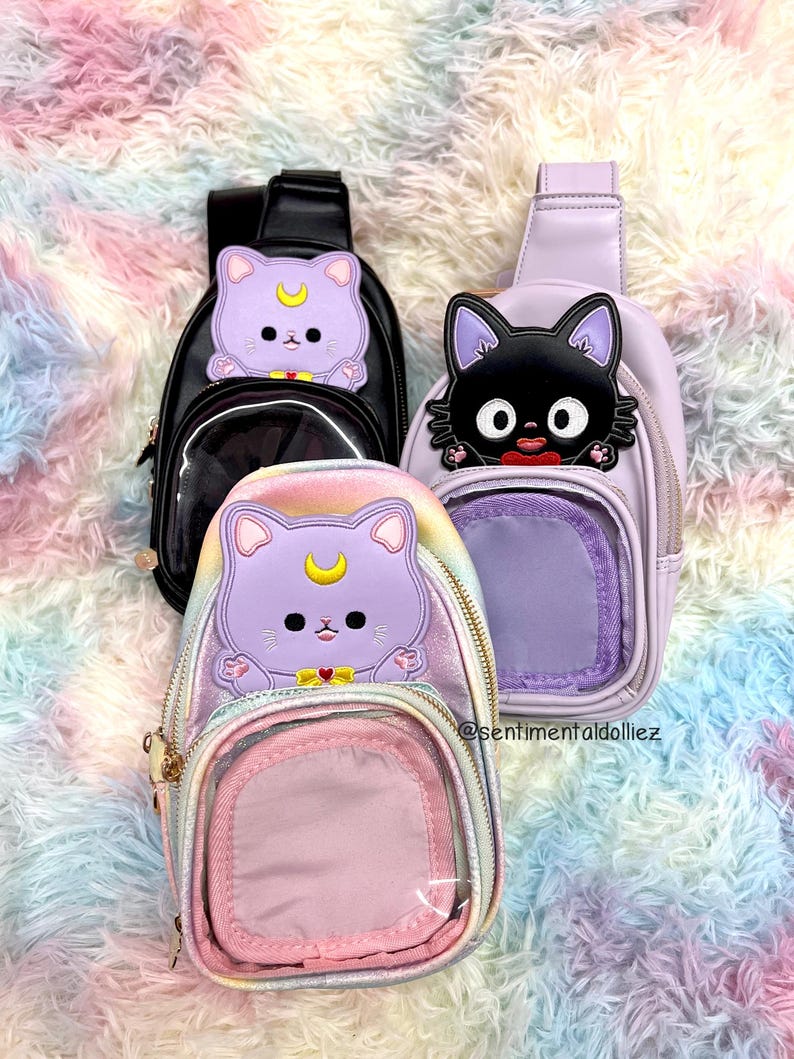 K&ouml;nnte beinhalten: Drei kleine, transparente, durchsichtige Umh&auml;ngetaschen mit Rei&szlig;verschluss. Die Taschen haben eine transparente Fronttasche und eine kleinere, undurchsichtige R&uuml;ckentasche. Die Taschen zeigen verschiedene Comicfiguren: eine schwarze Katze, eine lila Katze mit einem Halbmond und eine lila Katze mit einem Halbmond und einem Regenbogenhintergrund.