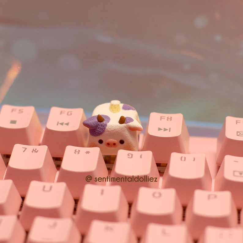 May include: A small, white cow-shaped keycap with purple spots sits on a pink keyboard. The keyboard has keys labeled with numbers and letters, including "F5", "F6", "F8", and "7&" . The text "@sentimentaldolliez" is printed on the keyboard.