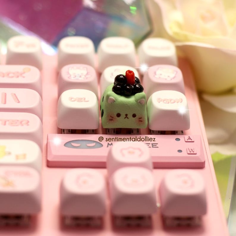 May include: A pink mechanical keyboard with custom keycaps. The keycaps are white and pink, some with cat designs. A green cat-shaped keycap with a boba tea design sits in the center. The text "@sentimentaldolliez" is on the keyboard.