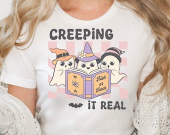 May include: White t-shirt with a Halloween graphic. The design shows three cartoon ghosts reading a book with the text "Trick or Teach" and the phrase "Creeping it Real". A bat graphic is below the text.