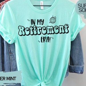 Op de afbeelding: Een heide mintgroen t-shirt met de tekst "In My Retirement Era!" in zwart, met een discobal-afbeelding boven de tekst. De tekst is in een retro-lettertype stijl.