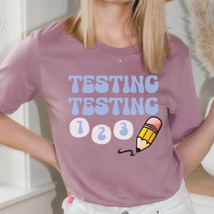 Puede incluir: Camiseta de color malva con las palabras "TESTING TESTING" en azul claro, los números "1 2 3" y un lápiz de dibujos animados. La camiseta tiene cuello redondo y mangas cortas.