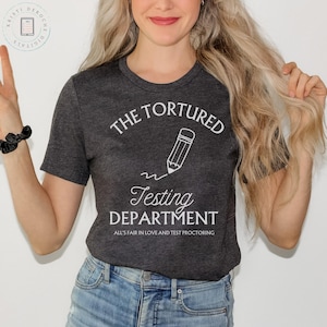 May include: A dark gray t-shirt with white text that reads "The Tortured Testing Department. All's fair in love and test proctoring." A white pencil with a squiggly line is above the word "Testing".