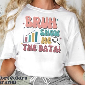 以下が含まれることがあります： 「Bruh Show Me The Data!」というレトロなフォントで書かれたグラフと虫眼鏡が描かれた白いTシャツ。テキストはピンク、ブルー、イエローです。