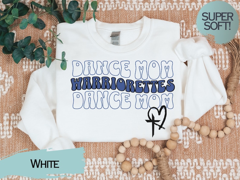 Puede incluir: Sudadera blanca con el texto "Dance Mom Warriorrettes Dance Mom" en fuente retro azul y blanca. Una cruz negra con un coraz&oacute;n est&aacute; en la esquina inferior derecha.