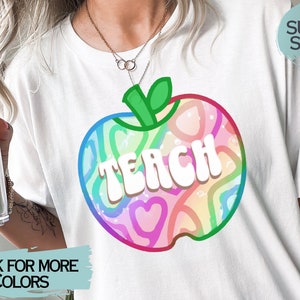 May include: White t-shirt with a colourful apple design and the word "TEACH" in white block letters. The apple design includes rainbow-coloured hearts and swirls. The shirt has a "SUPER SOFT!" label.