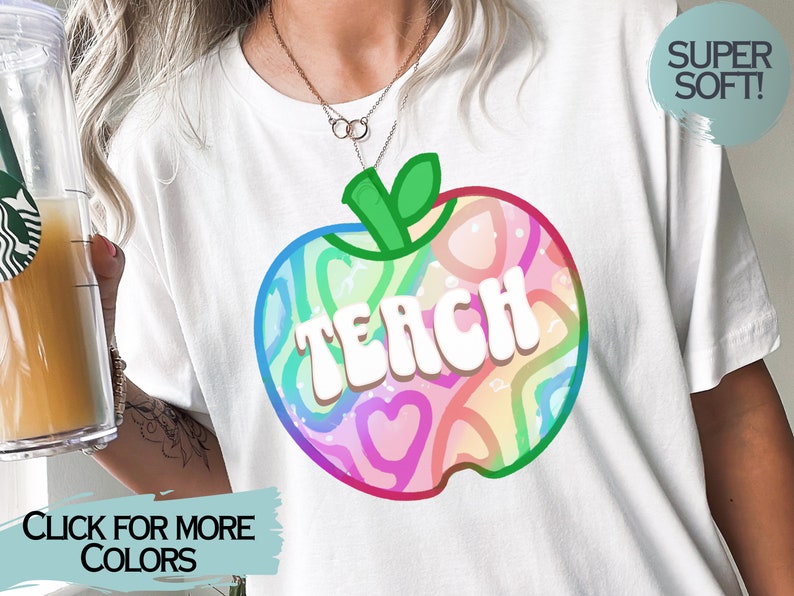 May include: White t-shirt with a colourful apple design and the word "TEACH" in white block letters. The apple design includes rainbow-coloured hearts and swirls. The shirt has a "SUPER SOFT!" label.