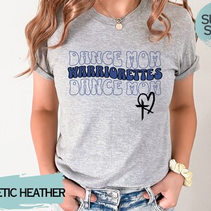 Puede incluir: Una camiseta gris jaspeada con el texto "Dance Mom Warriorrettes Dance Mom" en azul y negro con un dise&ntilde;o de cruz y coraz&oacute;n. La camiseta es s&uacute;per suave.