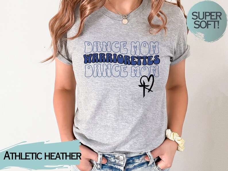 Puede incluir: Una camiseta gris jaspeada con el texto "Dance Mom Warriorrettes Dance Mom" en azul y negro con un dise&ntilde;o de cruz y coraz&oacute;n. La camiseta es s&uacute;per suave.