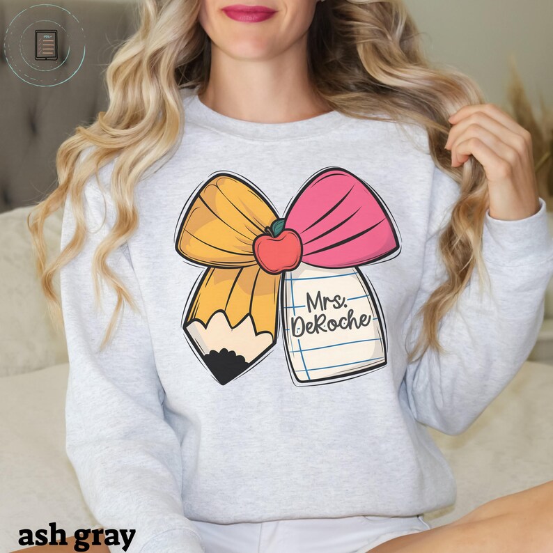 Puede incluir: Sudadera gris ceniza con un dise&ntilde;o de l&aacute;piz y lazo. El lazo es rosa y amarillo y tiene una manzana roja en el centro. El l&aacute;piz es amarillo y tiene una goma negra. El texto "Mrs. DeRoche" est&aacute; escrito en una libreta blanca.