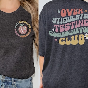 以下が含まれることがあります： 暗いグレーのTシャツ。前面には、ディスコボールのデザインのリンゴと「Over Stimulated Testing Coordinator」というレトロなグラフィックがプリントされています。背面には、星とデイジーがついたレトロなフォントで「Over Stimulated Testing Coordinators Club」という文字がプリントされています。