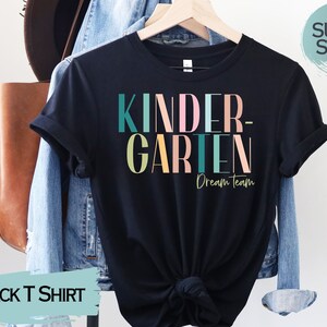 May include: Black t-shirt with the text "Kindergarten Dream Team" in colorful letters. The shirt is on a hanger and has a relaxed fit.