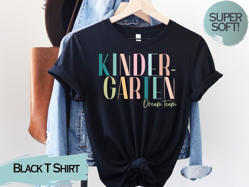 May include: Black t-shirt with the text "Kindergarten Dream Team" in colorful letters. The shirt is on a hanger and has a relaxed fit.