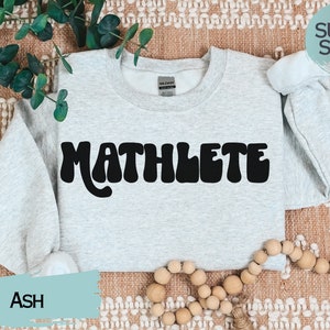 May include: A gray sweatshirt with the word "MATHLETE" printed in black retro-style lettering. The sweatshirt is on a brown and white patterned surface. The text "SUPER SOFT!" is in a speech bubble above the sweatshirt.