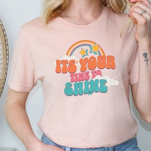Può includere: T-shirt rosa chiaro con un design retrò. La grafica include un arcobaleno, stelle e la scritta "IT'S YOUR TIME TO SHINE" in arancione, rosa e turchese. La maglietta è realizzata in materiale morbido.