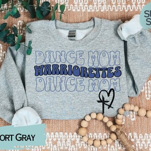 Puede incluir: Una sudadera gris con el texto "Dance Mom Warriorrettes Dance Mom" en letras azules y blancas. Una cruz con un coraz&oacute;n est&aacute; debajo del texto. La sudadera est&aacute; sobre una superficie con estampado marr&oacute;n y blanco.