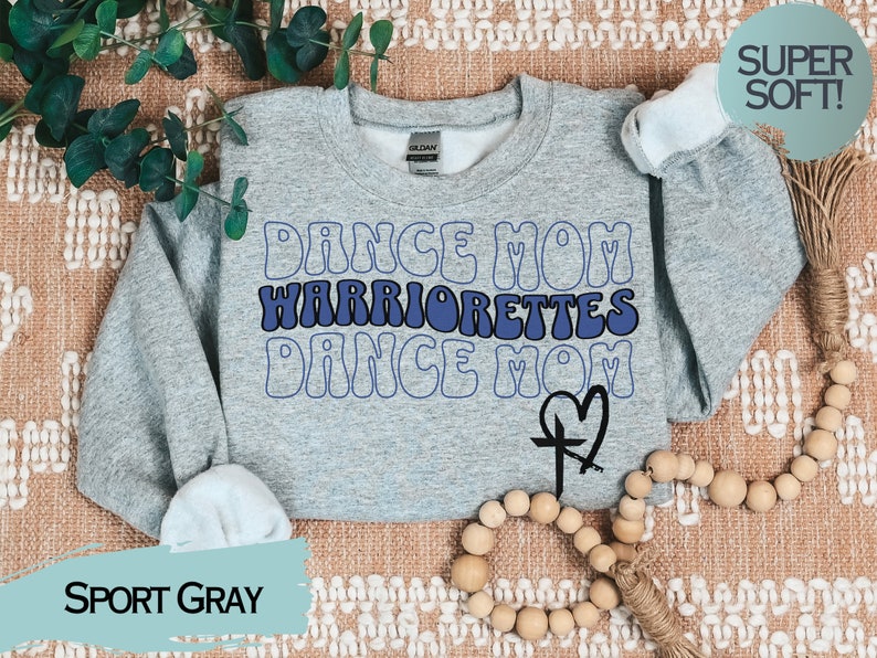 Puede incluir: Una sudadera gris con el texto "Dance Mom Warriorrettes Dance Mom" en letras azules y blancas. Una cruz con un coraz&oacute;n est&aacute; debajo del texto. La sudadera est&aacute; sobre una superficie con estampado marr&oacute;n y blanco.