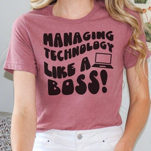 Puede incluir: Camiseta rosa polvoriento con el texto "MANAGING TECHNOLOGY LIKE A BOSS!" en negrita, negro, fuente de burbujas. Se incluye un gráfico de portátil negro. La camiseta es de cuello redondo y manga corta.