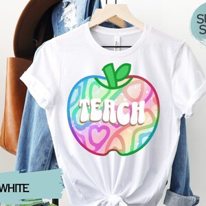 May include: White t-shirt with a colourful apple graphic. The apple design features the word "TEACH" in white, outlined in a rainbow of colours. The shirt is hanging on a wooden hanger. The words "SUPER SOFT!" are in a circle.