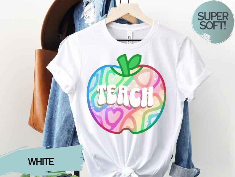 May include: White t-shirt with a colourful apple graphic. The apple design features the word "TEACH" in white, outlined in a rainbow of colours. The shirt is hanging on a wooden hanger. The words "SUPER SOFT!" are in a circle.