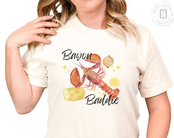 May include: A white t-shirt with a watercolor illustration of a crawfish, corn, lemon, and onion. The text "Bayou Baddie" is printed on the shirt.