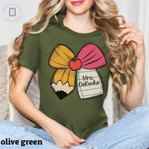 Puede incluir: Camiseta verde oliva con un lazo hecho de un l&aacute;piz y una cinta rosa. El lazo tiene una manzana roja en el centro y una nota de papel blanco que dice "Mrs. DeRoche".