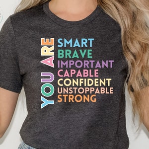 以下が含まれることがあります： 暗いグレーのTシャツに、「YOU ARE」という文字が緑、黄色、紫で書かれており、その後にさまざまな色の肯定的な言葉が並んでいます。SMART、BRAVE、IMPORTANT、CAPABLE、CONFIDENT、UNSTOPPABLE、STRONGです。