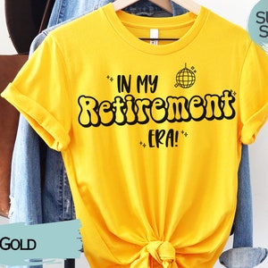 Op de afbeelding: Een geel T-shirt met de tekst "In My Retirement Era!" in zwarte bubbelletters. Een discobal-afbeelding bevindt zich boven de tekst. Het shirt hangt aan een hanger en heeft een "Super Soft!"-label.