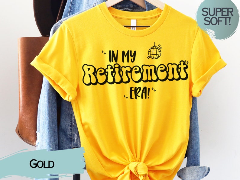 Op de afbeelding: Een geel T-shirt met de tekst "In My Retirement Era!" in zwarte bubbelletters. Een discobal-afbeelding bevindt zich boven de tekst. Het shirt hangt aan een hanger en heeft een "Super Soft!"-label.