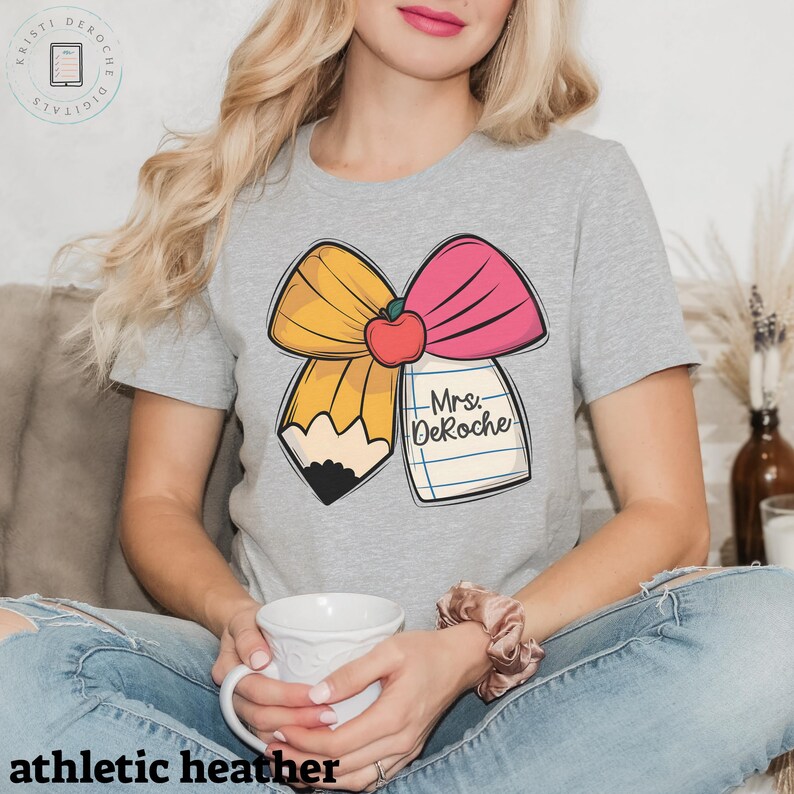 Puede incluir: Una camiseta gris con un lazo amarillo y rosa, una manzana roja y un l&aacute;piz. El l&aacute;piz est&aacute; unido a un trozo de papel con el texto "Mrs. DeRoche" escrito en &eacute;l.