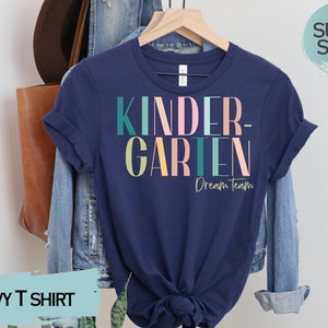 May include: Navy blue t-shirt with the text "KINDERGARTEN - Dream Team" in colorful letters. The shirt is made of a soft, comfortable material.
