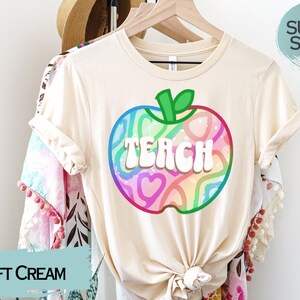 May include: A soft cream-coloured t-shirt with a colourful apple graphic. The apple design features the word "TEACH" in white, with a rainbow-coloured pattern inside. The shirt is hanging on a wooden hanger.