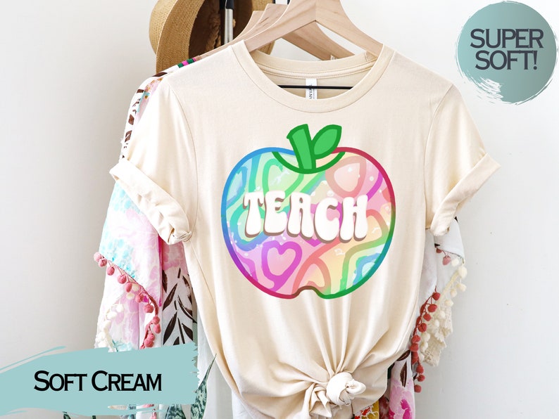 May include: A soft cream-coloured t-shirt with a colourful apple graphic. The apple design features the word "TEACH" in white, with a rainbow-coloured pattern inside. The shirt is hanging on a wooden hanger.