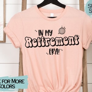 Op de afbeelding: Een roze T-shirt met de tekst "In My Retirement Era!" in zwart, met een discobal-afbeelding. De tekst is in een retro-lettertype.