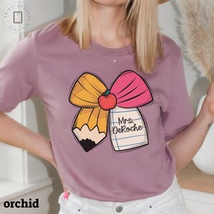 Puede incluir: Una camiseta morada con un gr&aacute;fico de un lazo amarillo y rosa con una manzana roja y un l&aacute;piz. El l&aacute;piz est&aacute; envuelto en un trozo de papel con el texto "Mrs. DeRoche" escrito en &eacute;l.