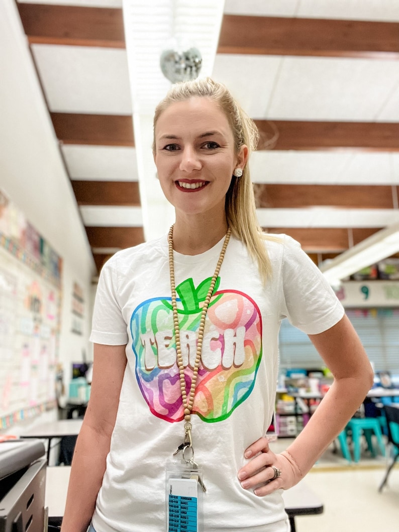 May include: White t-shirt with a colourful apple graphic and the word "TEACH" in large letters. The apple design features rainbow colours. A wooden bead necklace and a clear ID holder are also visible.