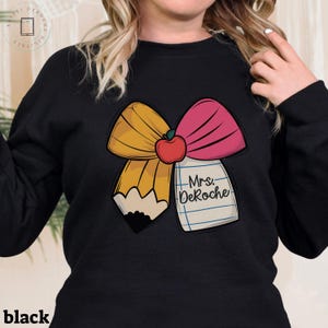 Puede incluir: Sudadera negra con un gr&aacute;fico de un lazo amarillo y rosa con una manzana roja en el centro. El lazo est&aacute; dise&ntilde;ado para parecer un l&aacute;piz con un bloc de notas que dice "Mrs. DeRoche".