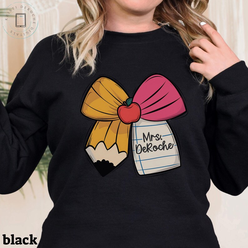 Puede incluir: Sudadera negra con un gr&aacute;fico de un lazo amarillo y rosa con una manzana roja en el centro. El lazo est&aacute; dise&ntilde;ado para parecer un l&aacute;piz con un bloc de notas que dice "Mrs. DeRoche".