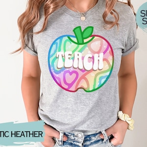 May include: A grey athletic heather t-shirt with a colourful apple design and the word "TEACH" in white. The apple design features a rainbow of colours and heart shapes. The shirt is labelled "SUPER SOFT!"
