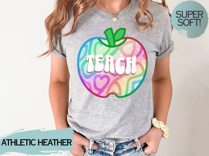 May include: A grey athletic heather t-shirt with a colourful apple design and the word "TEACH" in white. The apple design features a rainbow of colours and heart shapes. The shirt is labelled "SUPER SOFT!"