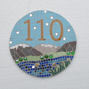 Peut inclure: Panneau de numéro de maison en mosaïque circulaire avec le numéro "110" en marron sur fond bleu. Le design comprend un paysage avec des montagnes, une rivière et des détails verts. La mosaïque est faite de carreaux de différentes couleurs.